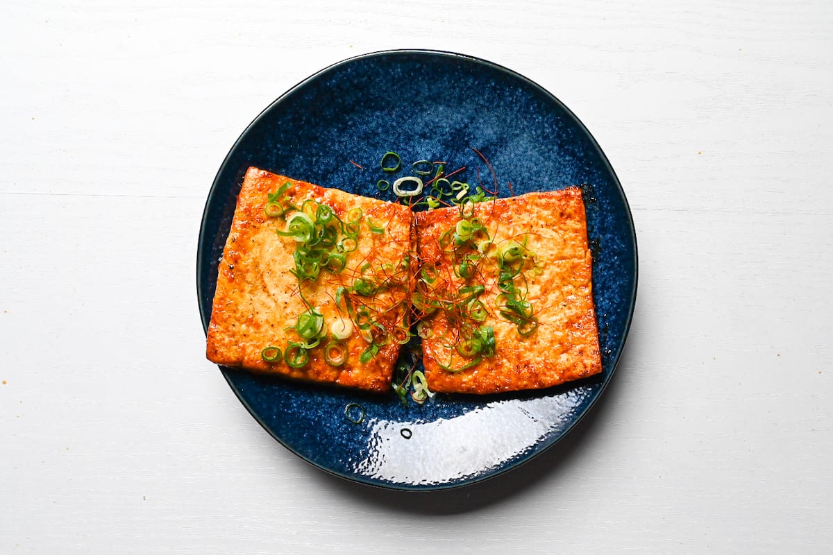 Two tofu steaks on a blue plate garnished with spring onion and red chili threads