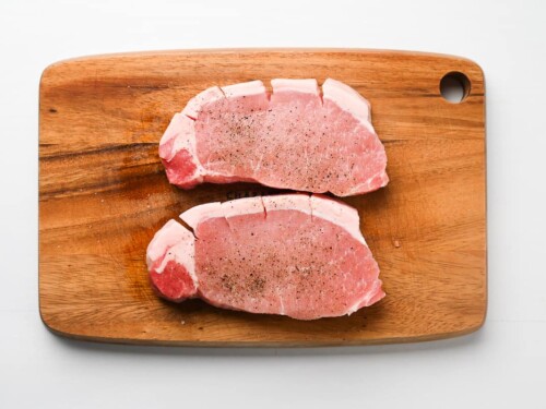 2 pork chops on a chopping board sprinkled with salt and pepper