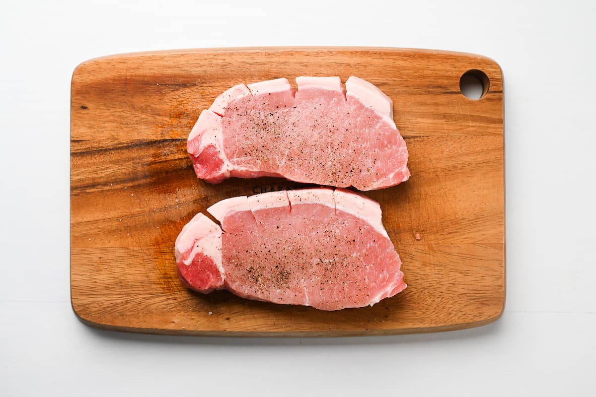 2 pork chops on a chopping board sprinkled with salt and pepper