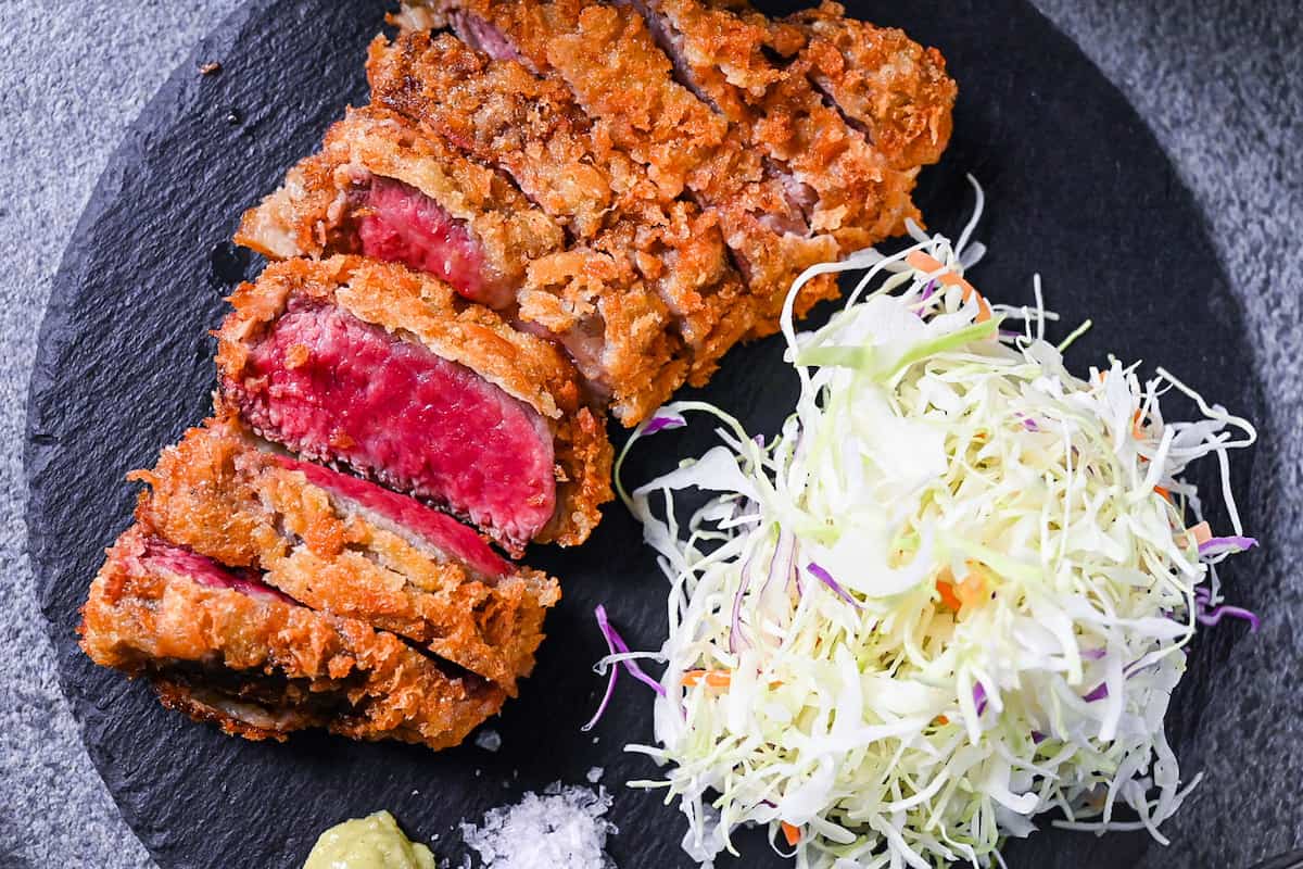 Gyukatsu (Japanese beef steak cutlet) served with cabbage, salt flakes and wasabi on a black slate plate