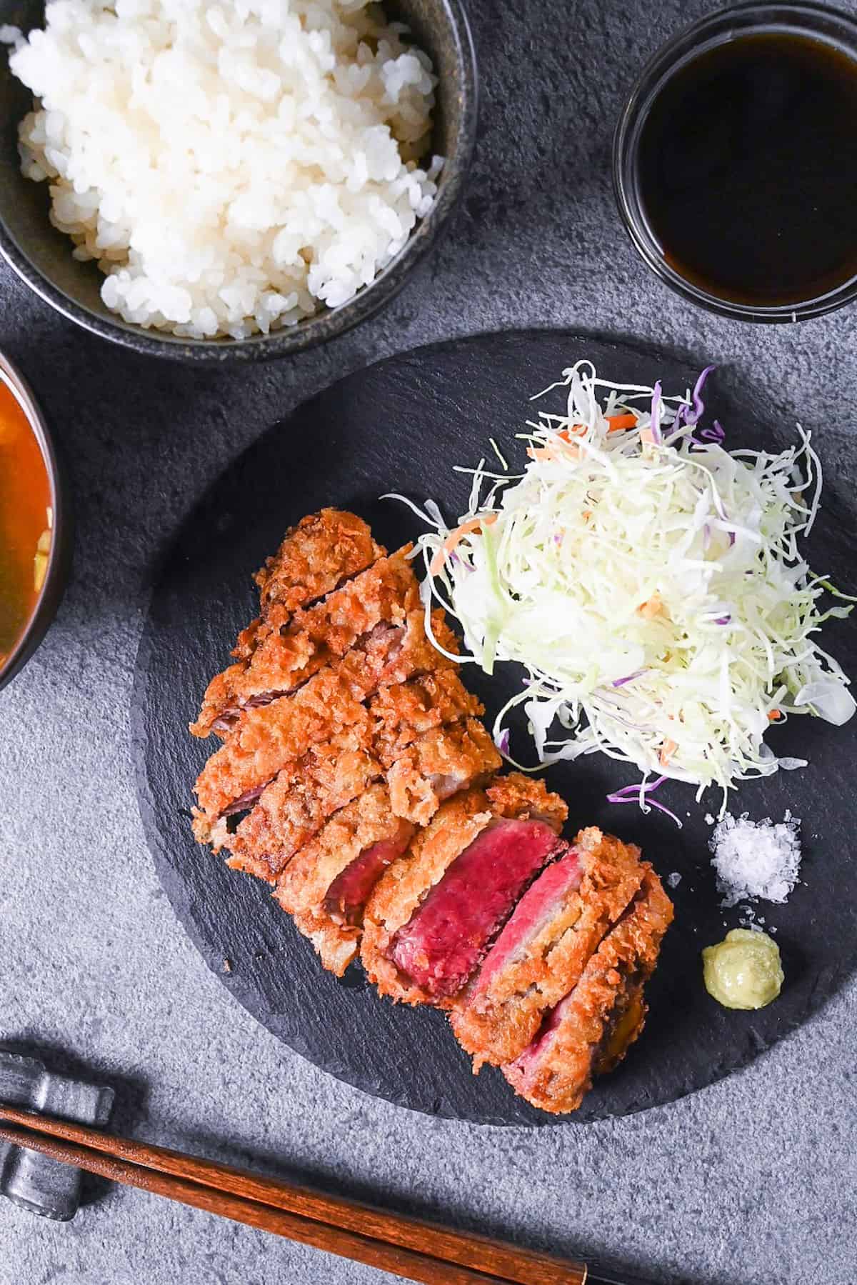 Gyukatsu (Japanese beef steak cutlet) served with cabbage, salt flakes and wasabi on a black slate plate