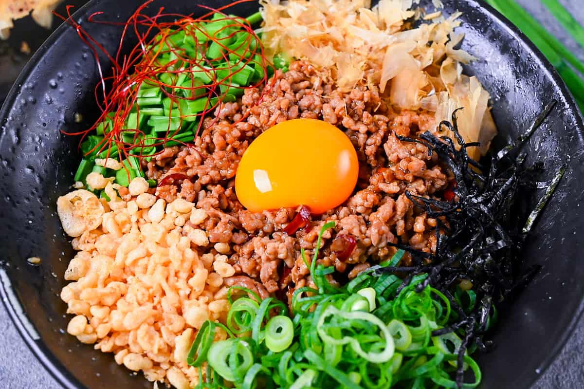 Taiwan Mazesoba served in a black ramen bowl and topped with seasoned pork mince, spring onions, Chinese chives, katsuobushi, tenkasu, kizami nori and a raw egg yolk