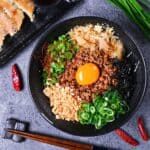 Taiwan Mazesoba served in a black ramen bowl and topped with seasoned pork mince, spring onions, Chinese chives, katsuobushi, tenkasu, kizami nori and a raw egg yolk