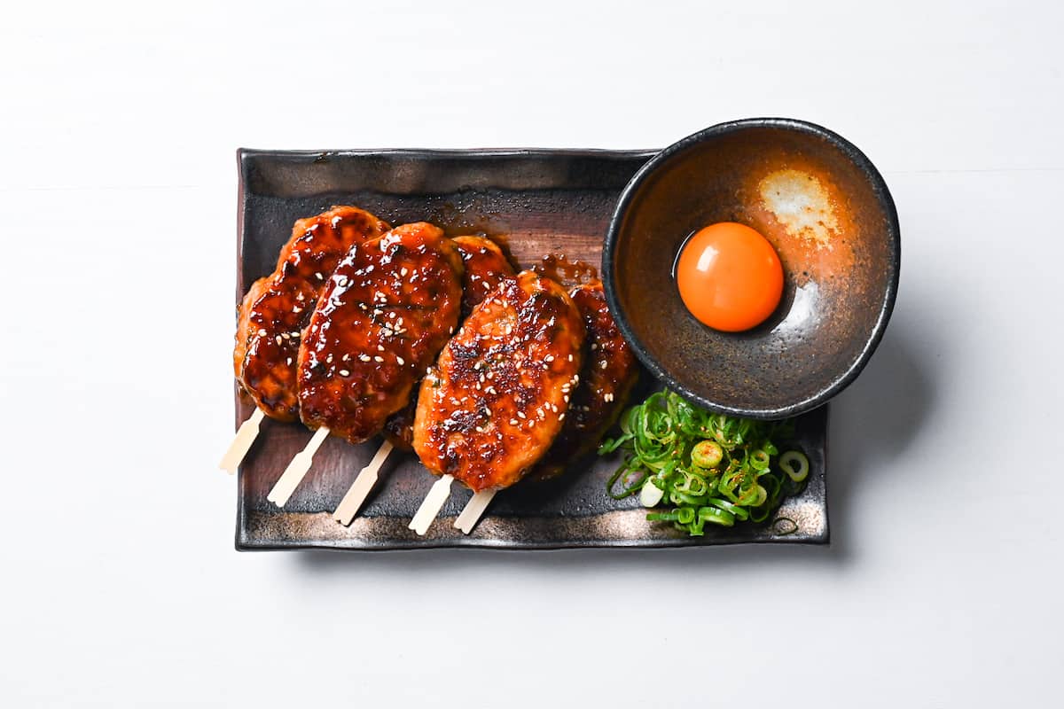 5 hand shaped chicken tsukune meatballs served on a rectangular plate with spring onion and raw egg yolk for dipping