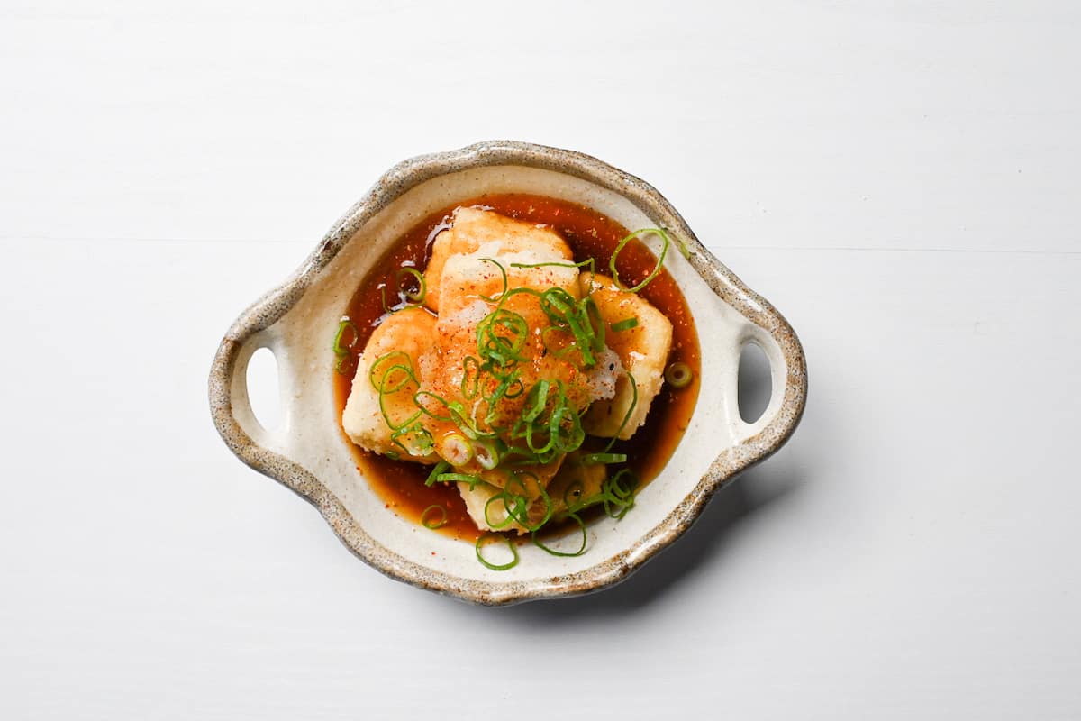 Agedashi tofu topped with chopped spring onion, grated daikon and shichimi pepper in a cream coloured serving bowl with handles