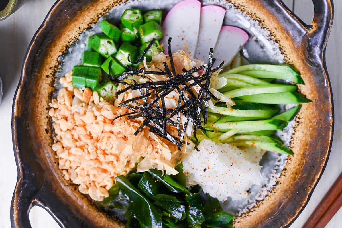 Hiyashi tanuki udon topped with tenkasu, okra, wakame, grated daikon, kamaboko, cucumber, katsuobushi and kizami nori top down view close up