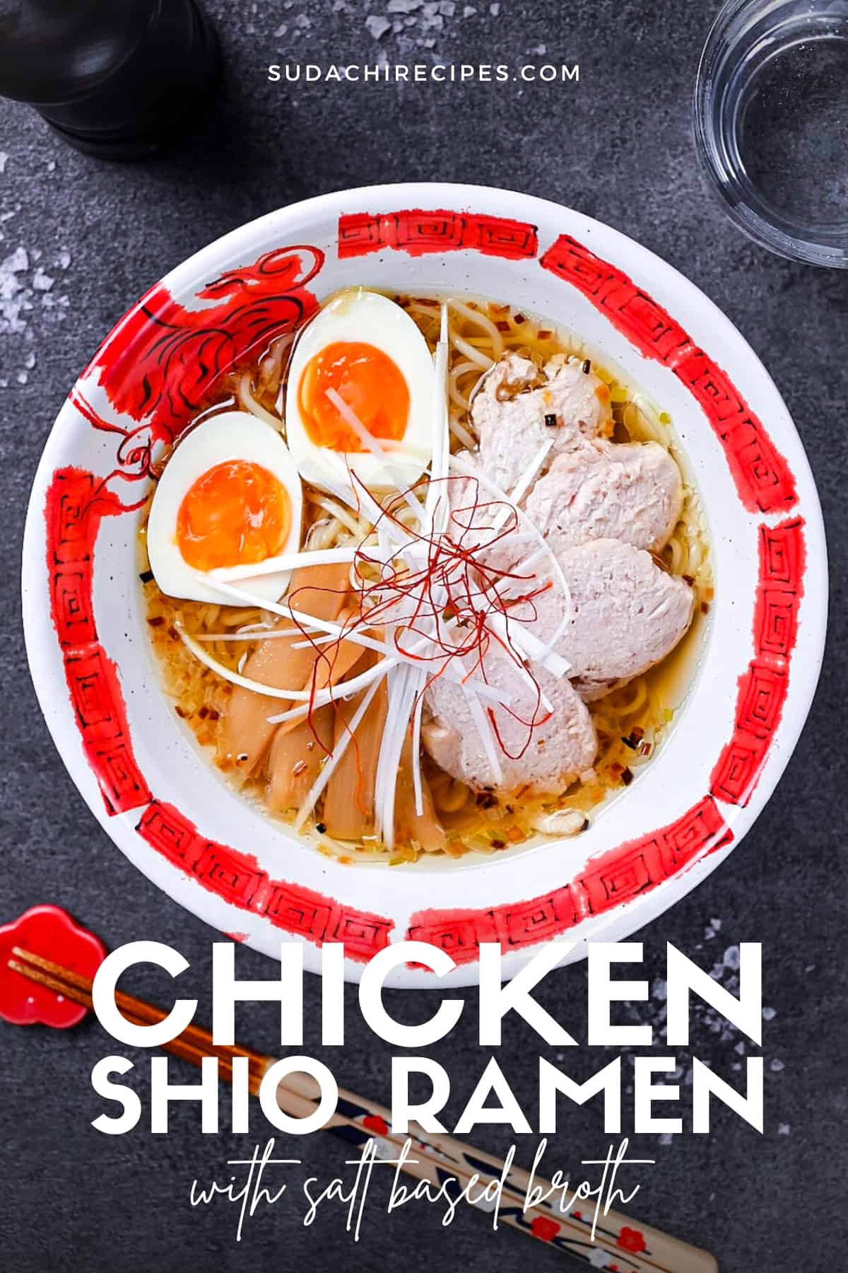 Japanese shio ramen topped with slices of chicken, menma (bamboo shoots), soft boiled egg, white spring onion and chilli threads in a red and white ramen bowl on a grey background
