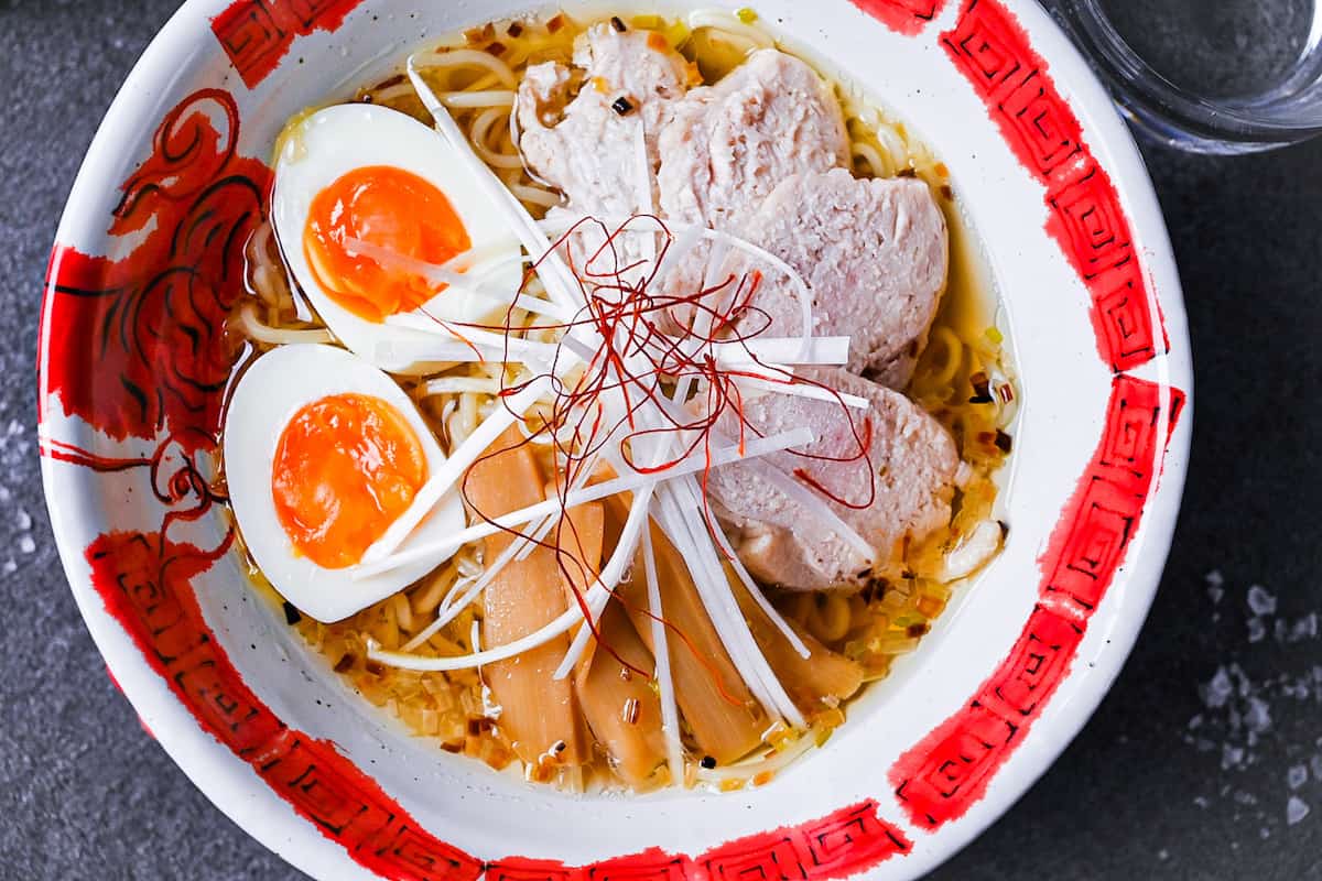 Japanese shio ramen topped with slices of chicken, menma (bamboo shoots), soft boiled egg, white spring onion and chilli threads in a red and white ramen bowl on a grey background