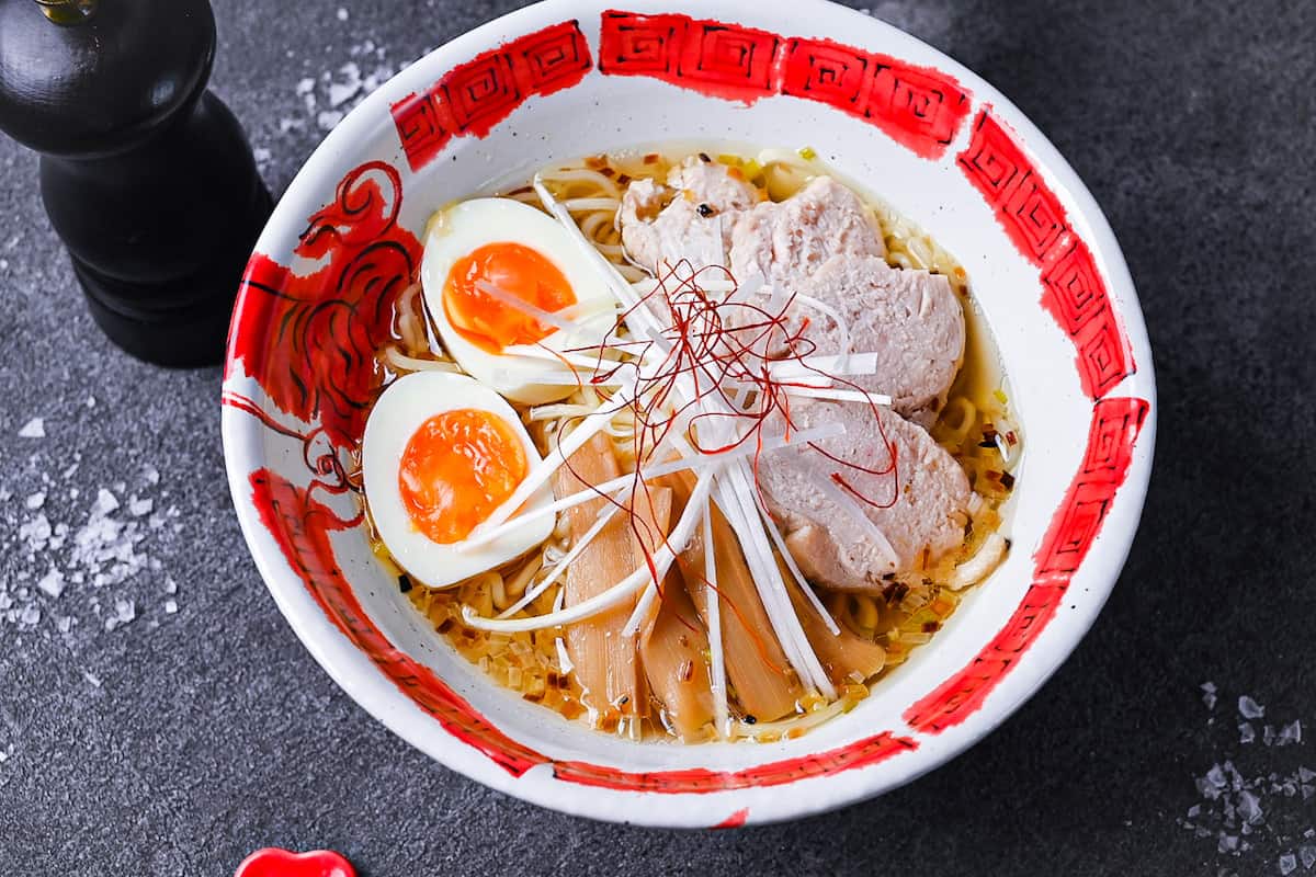 Japanese shio ramen topped with slices of chicken, menma (bamboo shoots), soft boiled egg, white spring onion and chilli threads in a red and white ramen bowl on a grey background