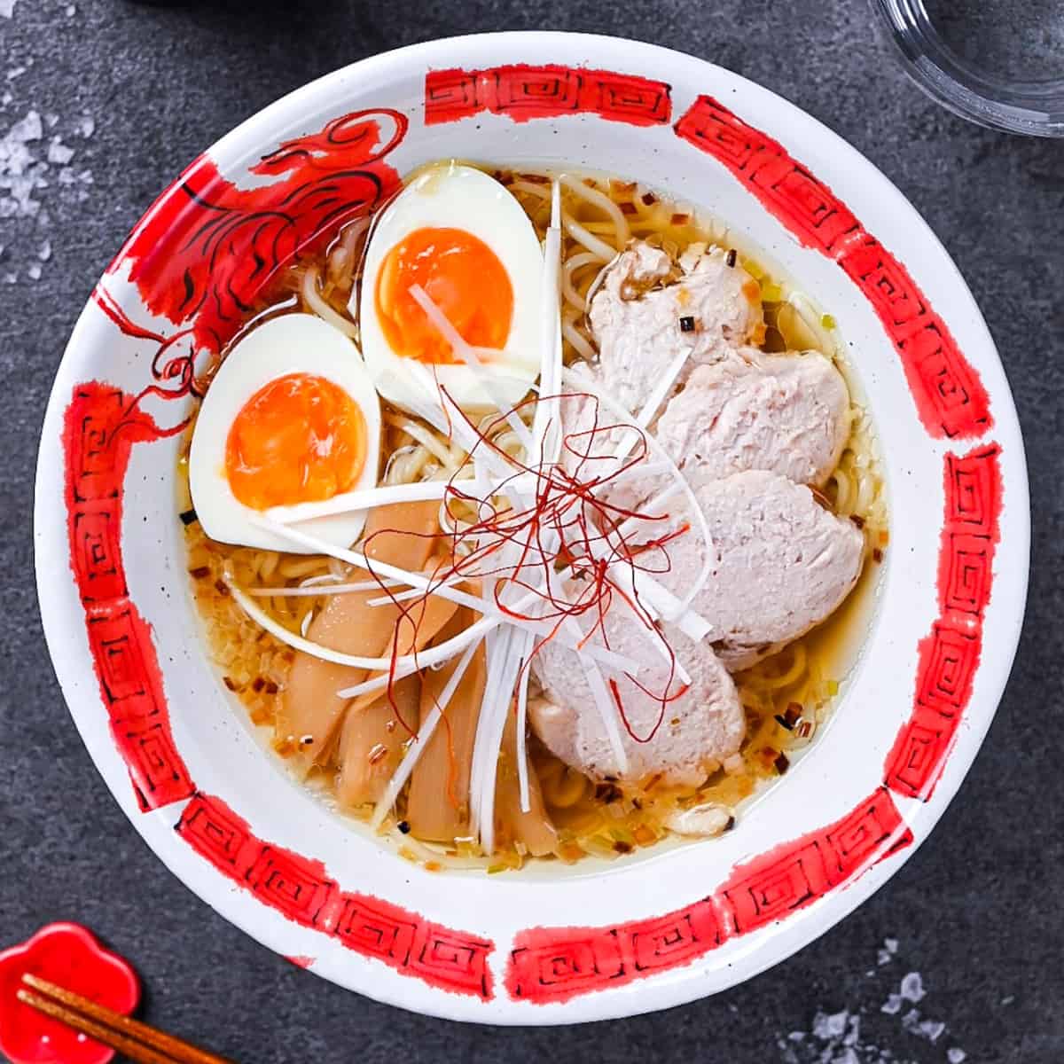 Japanese shio ramen topped with slices of chicken, menma (bamboo shoots), soft boiled egg, white spring onion and chilli threads in a red and white ramen bowl on a grey background