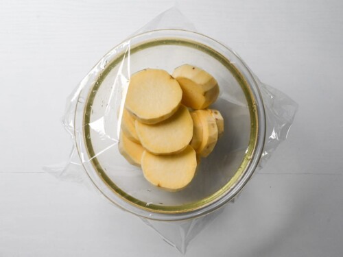 Slices of peeled sweet potato in a glass bowl with plastic wrap over the top