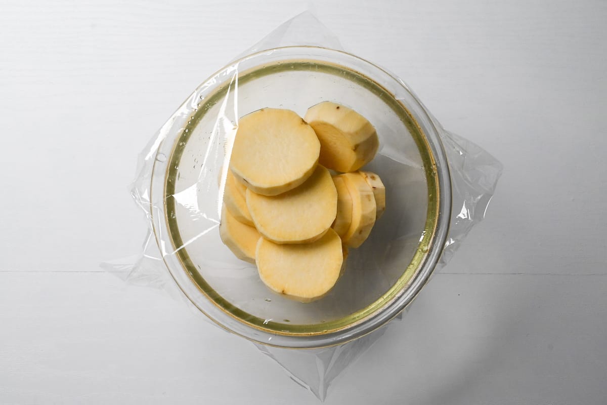 Slices of peeled sweet potato in a glass bowl with plastic wrap over the top