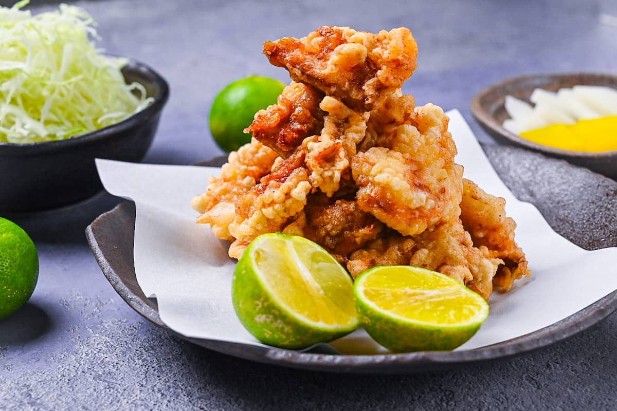 toriten (chicken tempura) piled up on a brown plate with two halves of kabosu citrus with shredded cabbage in the background