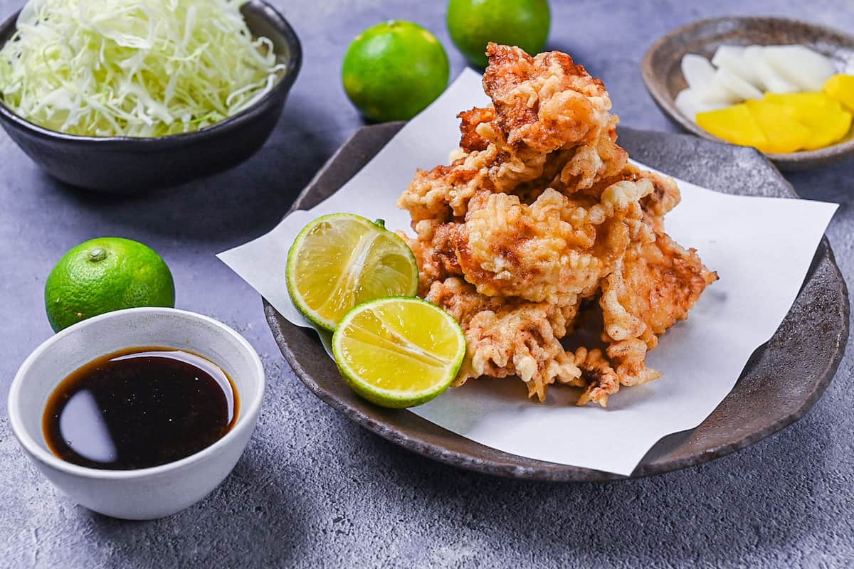 toriten (chicken tempura) piled up on a brown plate with two halves of kabosu citrus and dipping sauce on the side