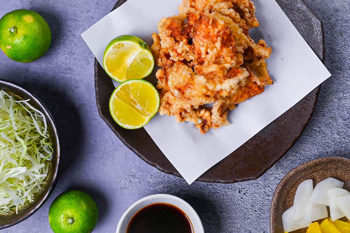 toriten (chicken tempura) piled up on a brown plate with two halves of kabosu citrus next to a bowl of shredded cabbage, daikon pickles and a dish of dipping sauce