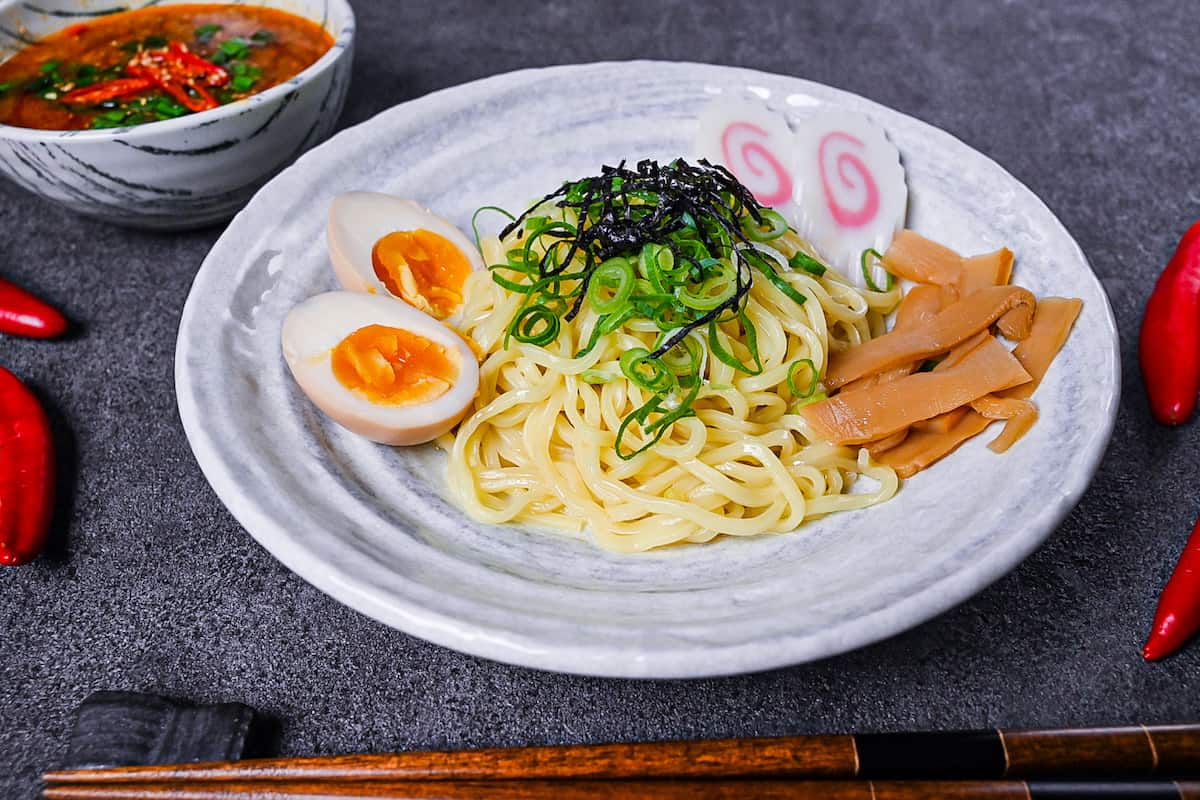 Japanese ramen noodles on a white plate with menma, narutomaki and ramen eggs served next to a bowl of spicy tsukemen soup
