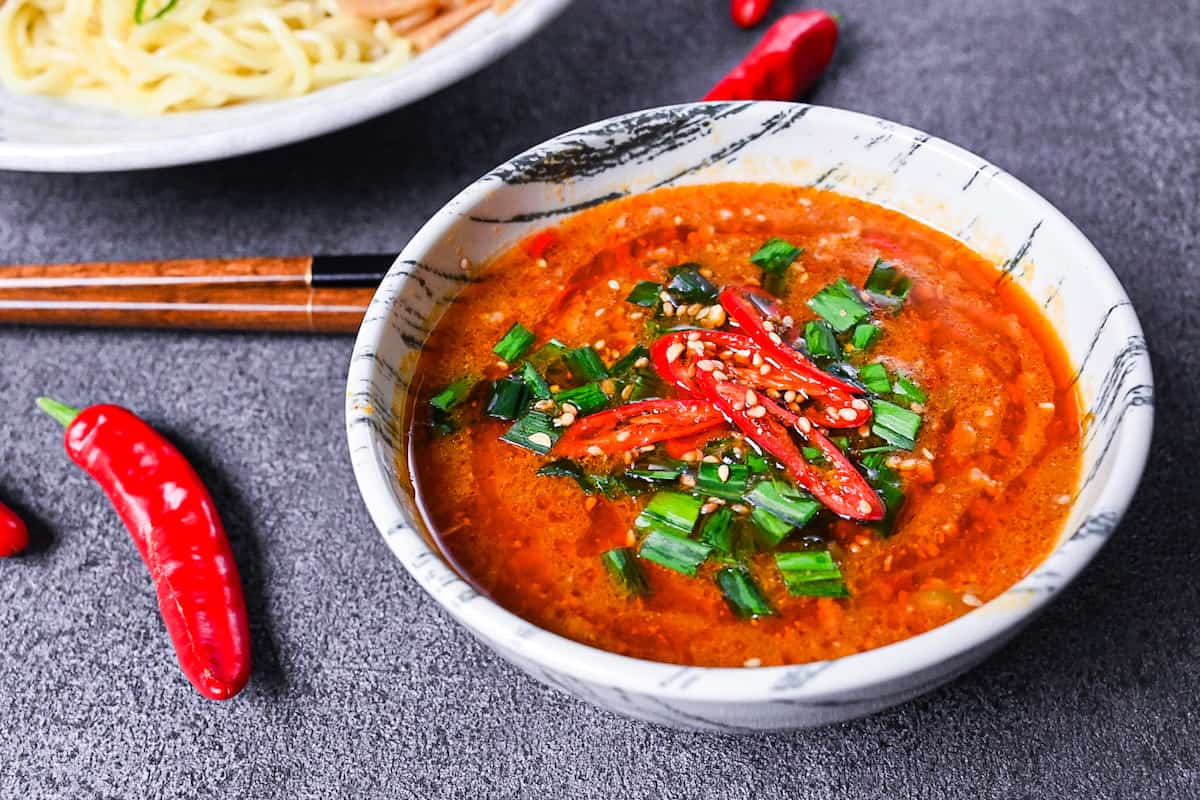 Spicy tsukemen soup in a black and white bowl topped with chives and fresh chili