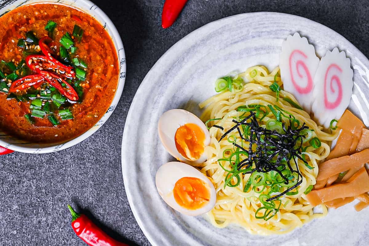 Japanese ramen noodles on a white plate with menma, narutomaki and ramen eggs served next to a bowl of spicy tsukemen soup top down