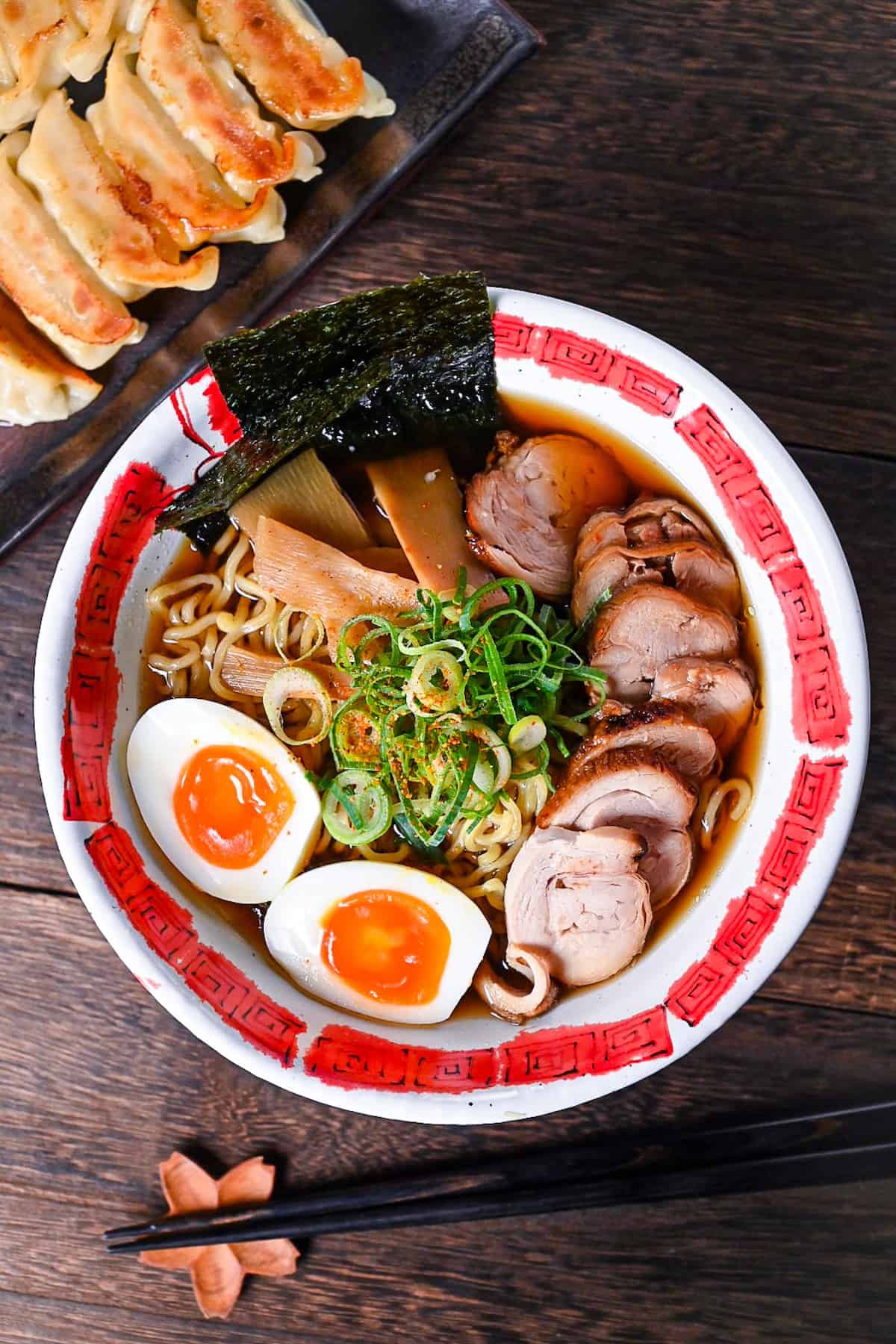 Homemade chicken chashu served on top of shoyu ramen in a white and red bowl next to a plate of gyoza