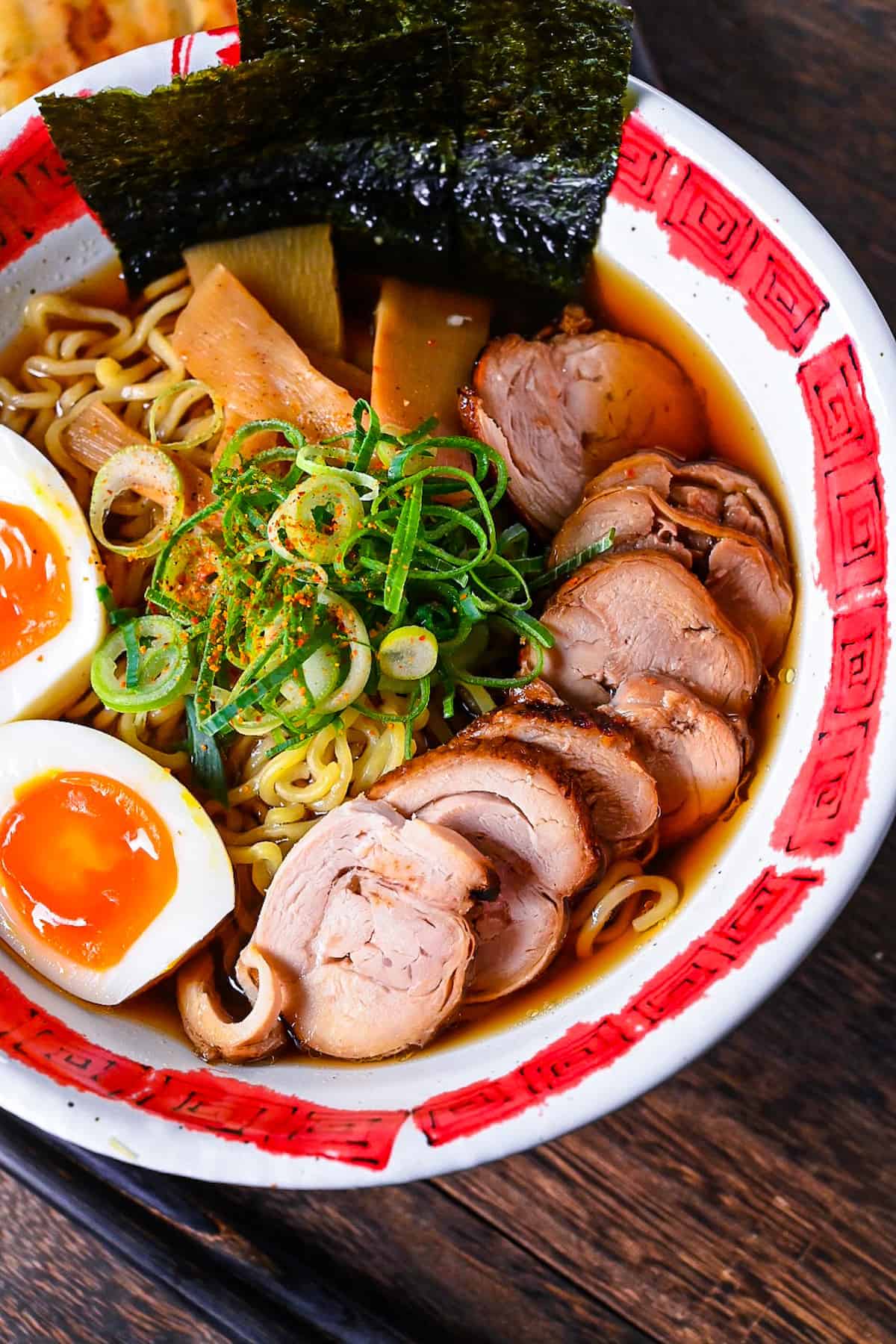 Homemade chicken chashu served on top of shoyu ramen in a white and red bowl next to a plate of gyoza