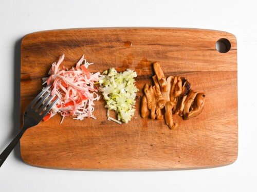 shredded imitation crab sticks, finely chopped spring onion and sliced shiitake mushrooms on a wooden chopping board