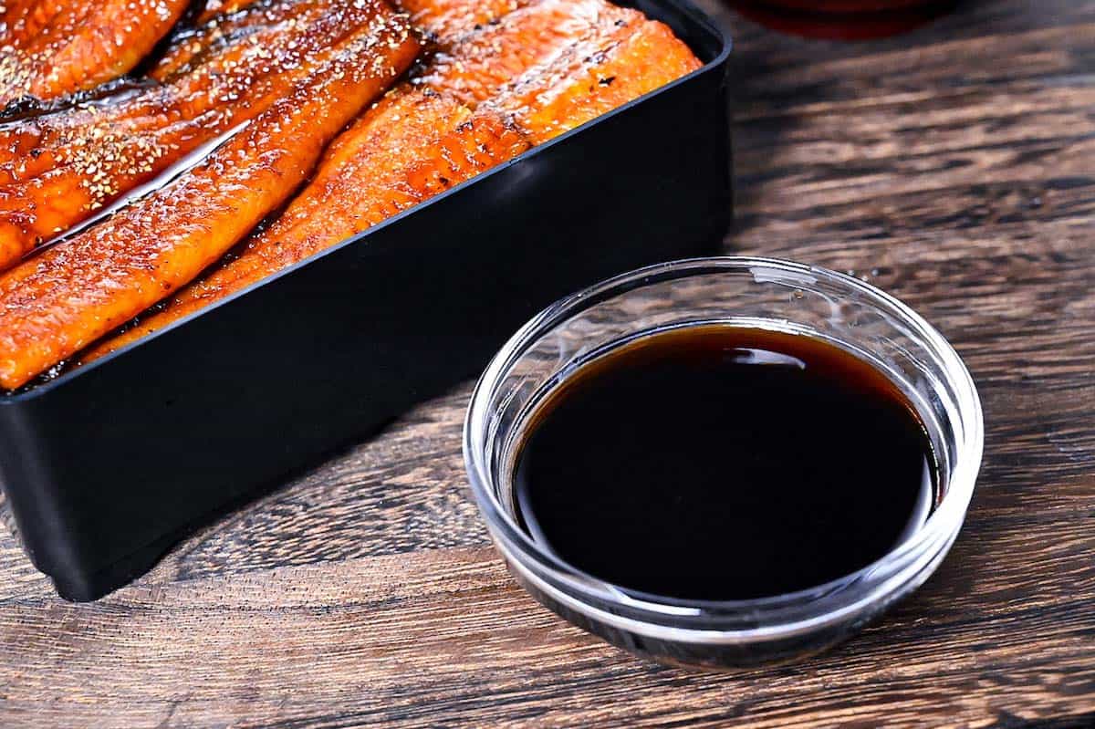 Unagi sauce in a small glass bowl next to grilled eel in a lacquerware box (unajyu)