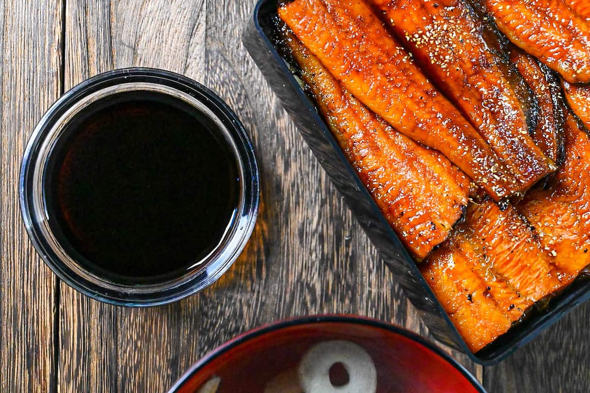 Unagi sauce in a small glass bowl next to grilled eel in a lacquerware box (unajyu) top down