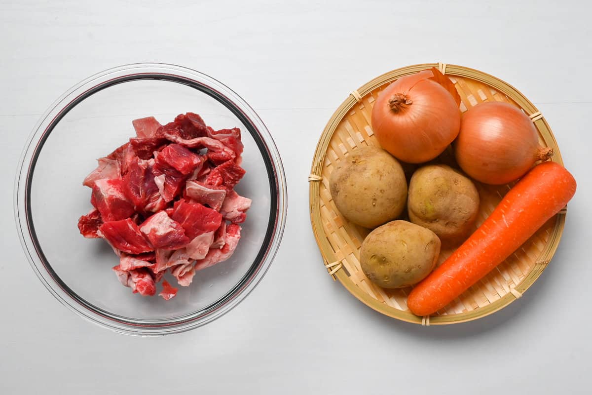Cubes of beef in a glass bowl and whole potatoes, onions and one carrot on a circular bamboo tray