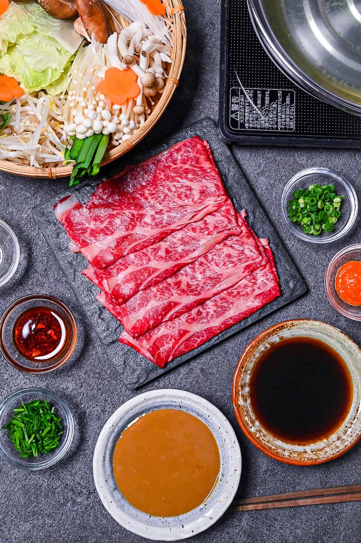 Ingredients to make shabu shabu on a gray background (a basket of vegetables and noodles, a plate of thinly sliced wagyu beef, various bowls of condiments and a bowl of sesame sauce and ponzu sauce)