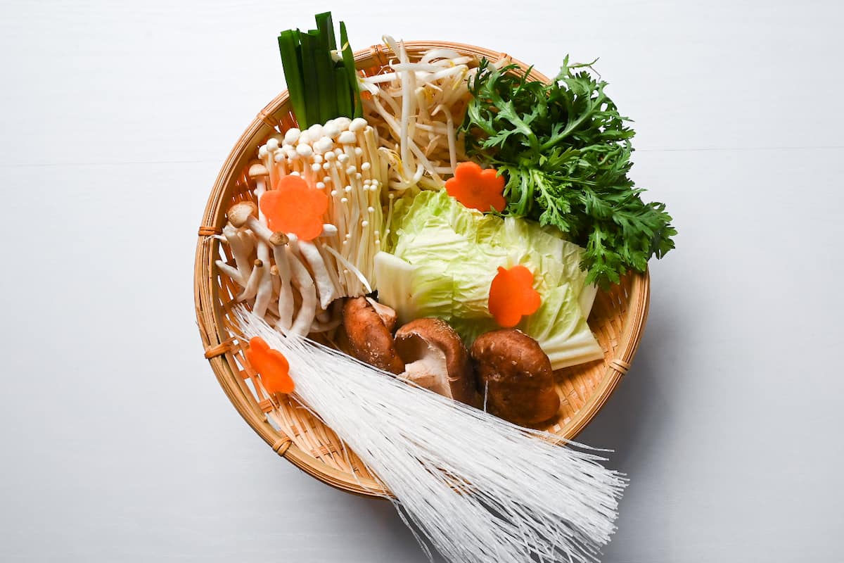 Vegetables and noodles for shabu shabu in a bamboo basket (chives, beansprouts, shungiku, napa cabbage, shimeji mushrooms, enoki mushrooms, carrots, shiitake mushrooms and glass noodles.)