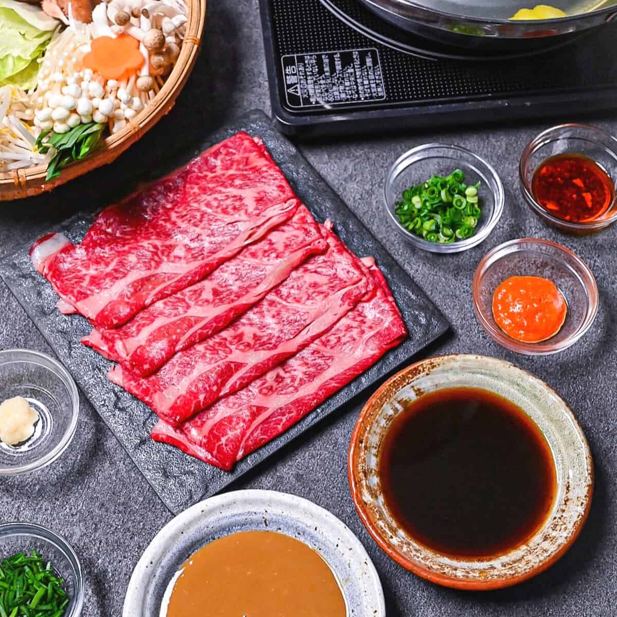 Ingredients to make shabu shabu on a gray background (a basket of vegetables and noodles, a plate of thinly sliced wagyu beef, various bowls of condiments and a bowl of sesame sauce and ponzu sauce)