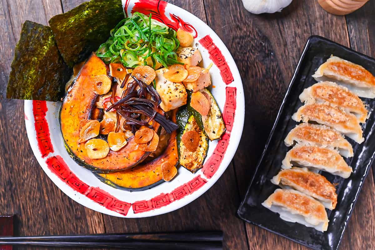 Spicy vegetarian miso ramen topped with oven baked kabocha, tofu and zucchini in a red and white bowl next to a plate of gyoza