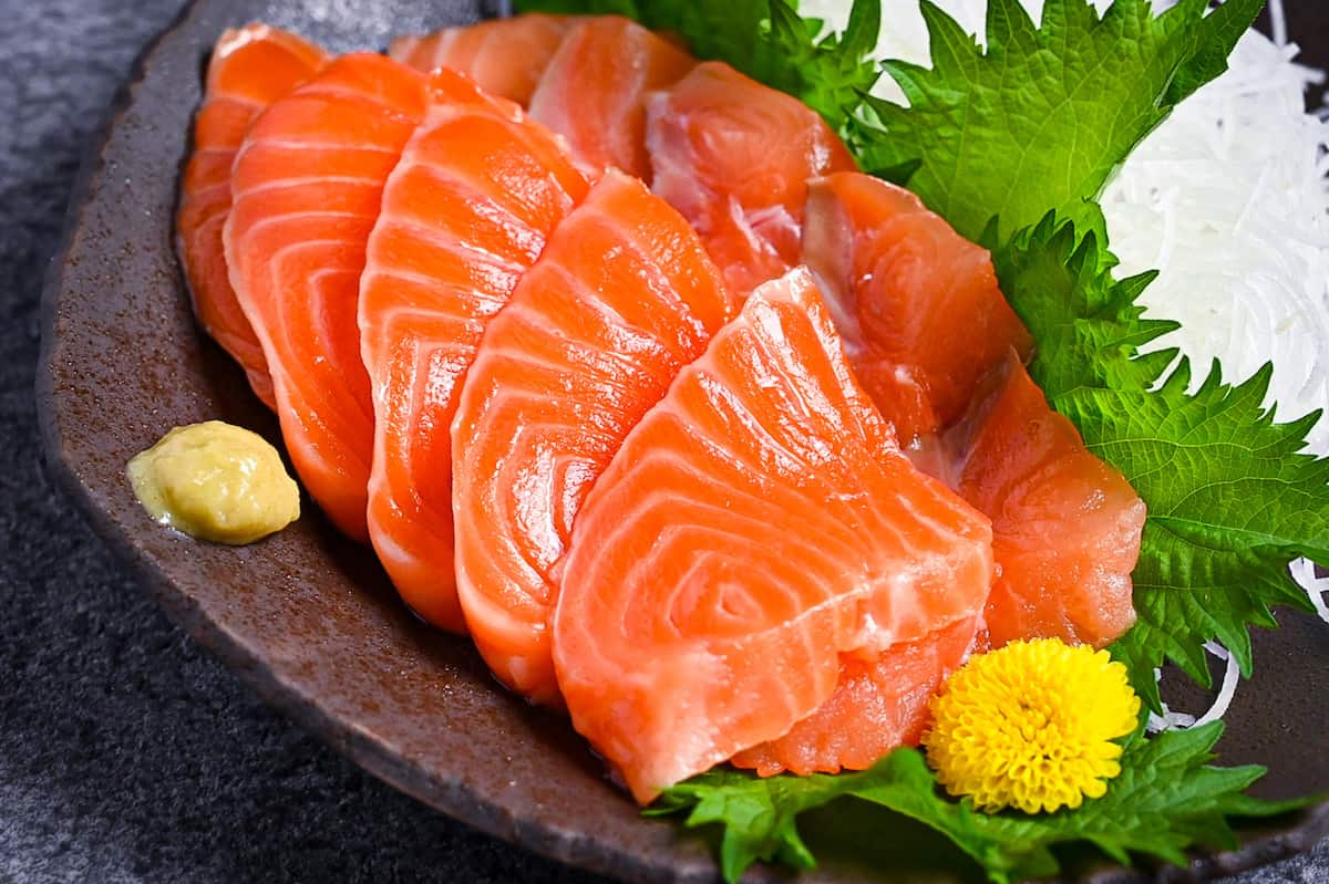 Slices of salmon sashimi on a brown plate with shredded daikon, shiso leaves and wasabi close up