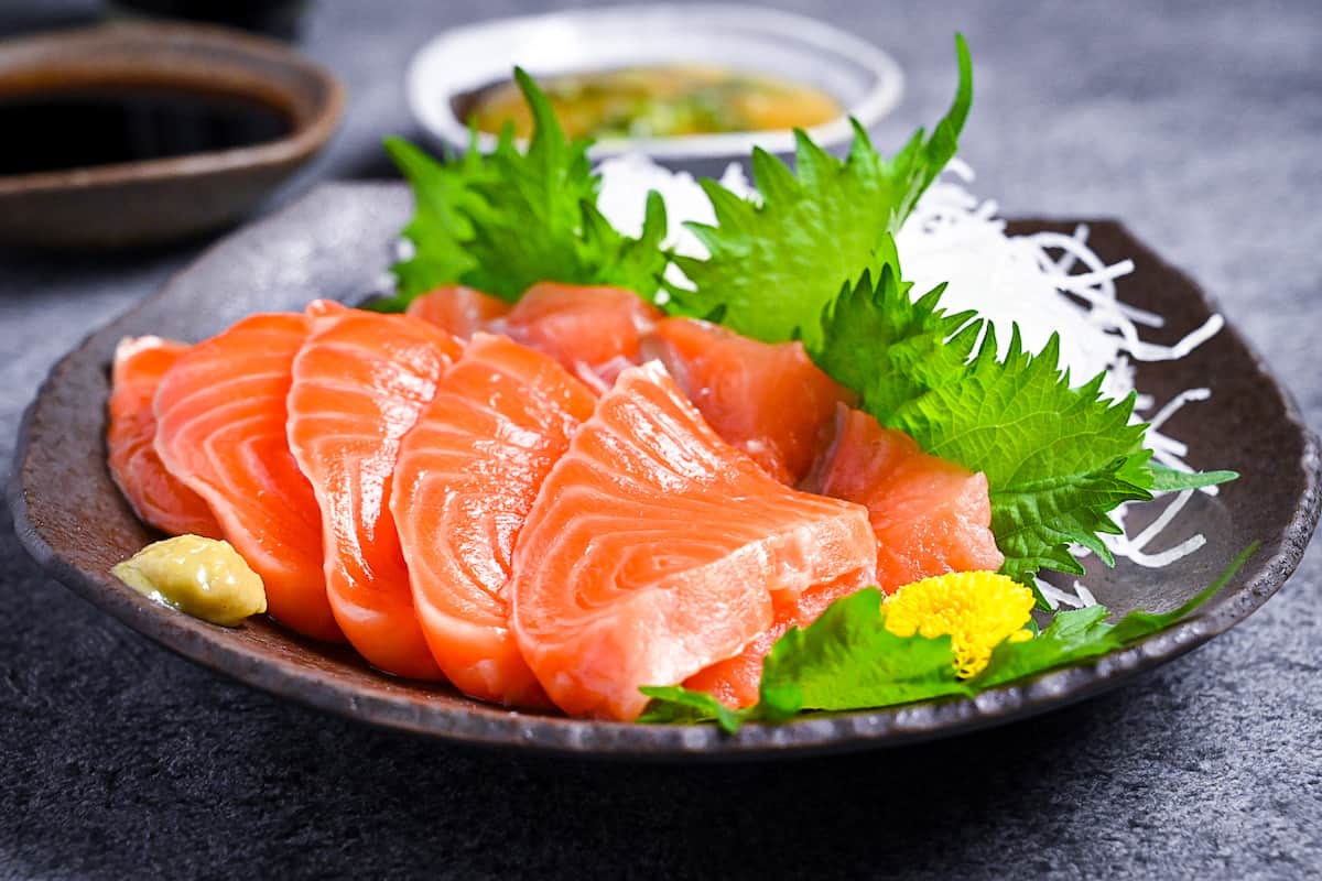 Slices of salmon sashimi on a brown plate with shredded daikon, shiso leaves and wasabi served with 3 types of sauce