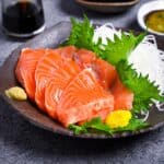 Slices of salmon sashimi on a brown plate with shredded daikon, shiso leaves and wasabi served with 3 types of sauce