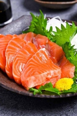 Slices of salmon sashimi on a brown plate with shredded daikon, shiso leaves and wasabi served with 3 types of sauce