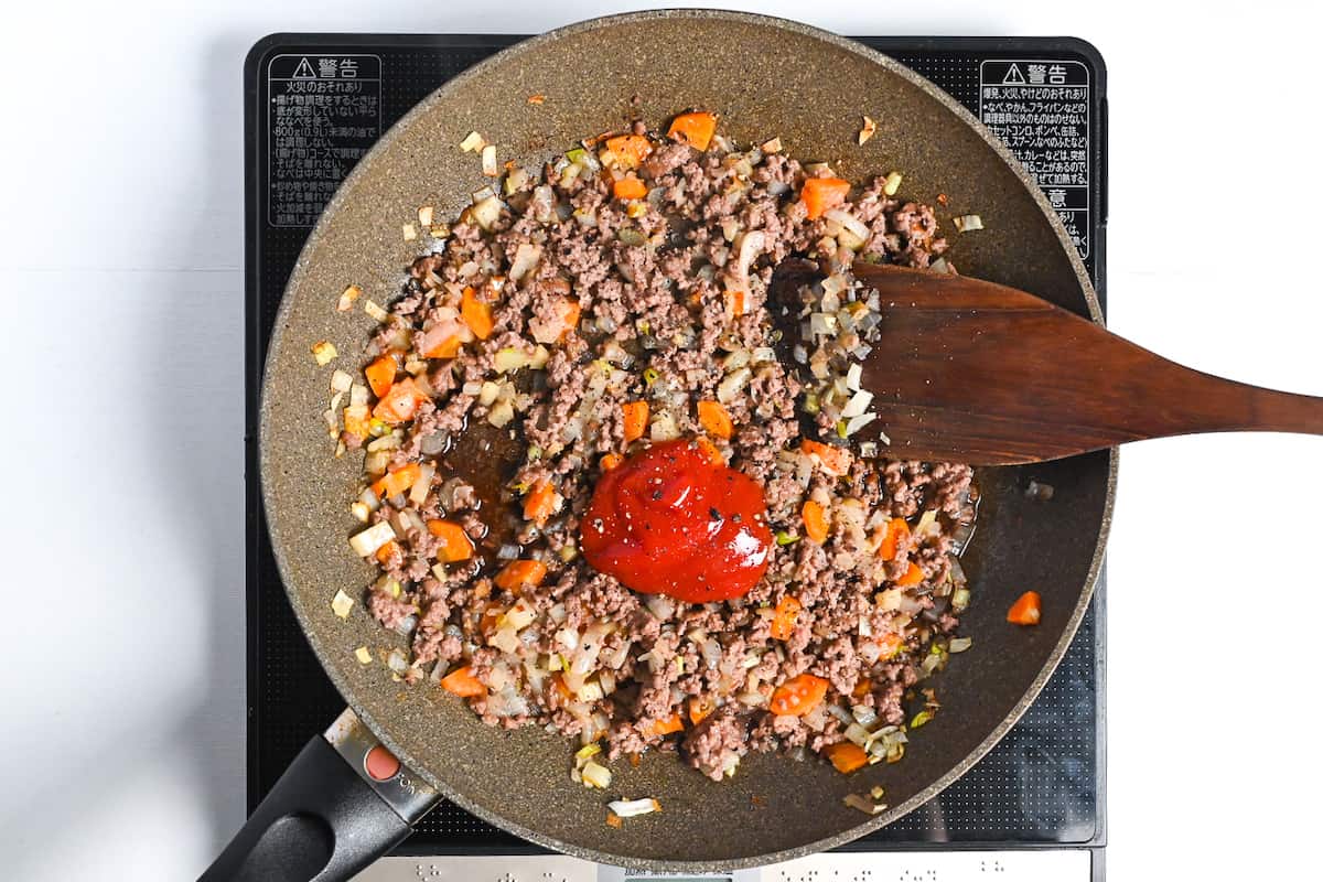 Ketchup and condiments added to cooked ground beef and vegetables in a large frying pan