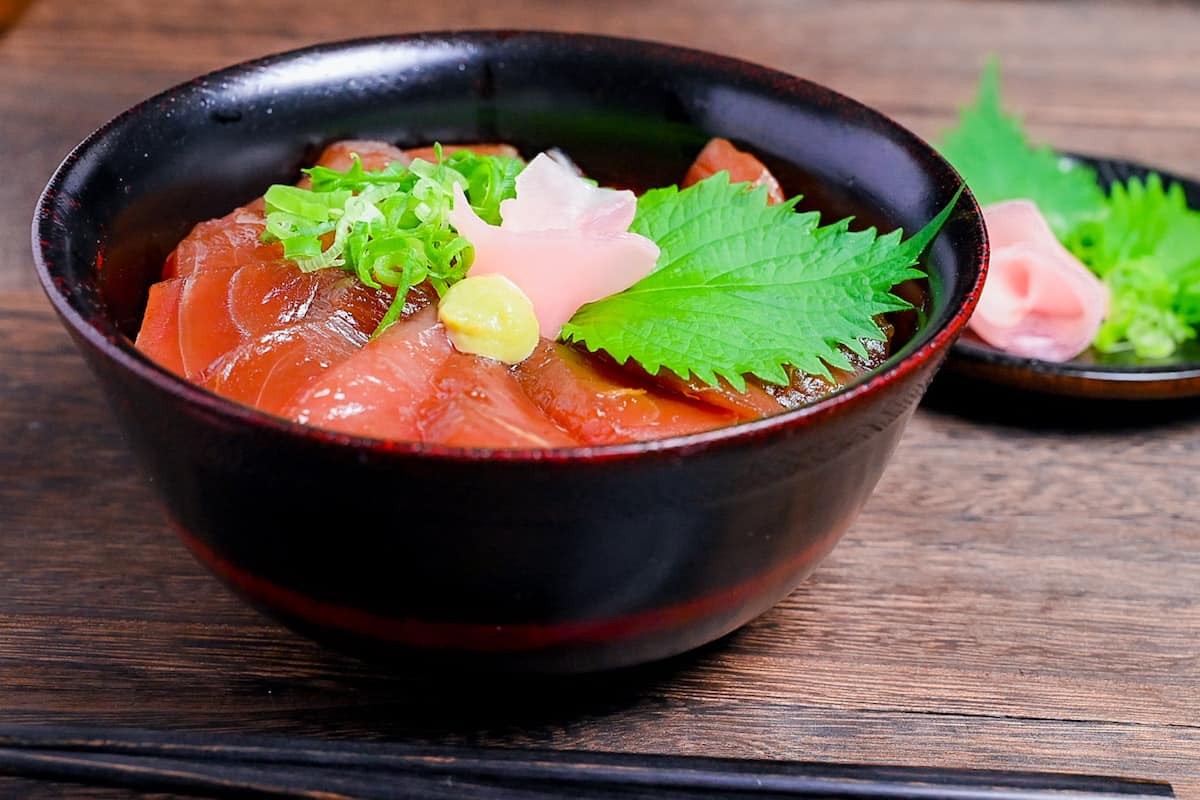 Tekkadon - vinegared rice topped with slices of fresh tuna sashimi, pickled ginger, wasabi, shiso and chopped spring onion in a black lacquerware bowl side view