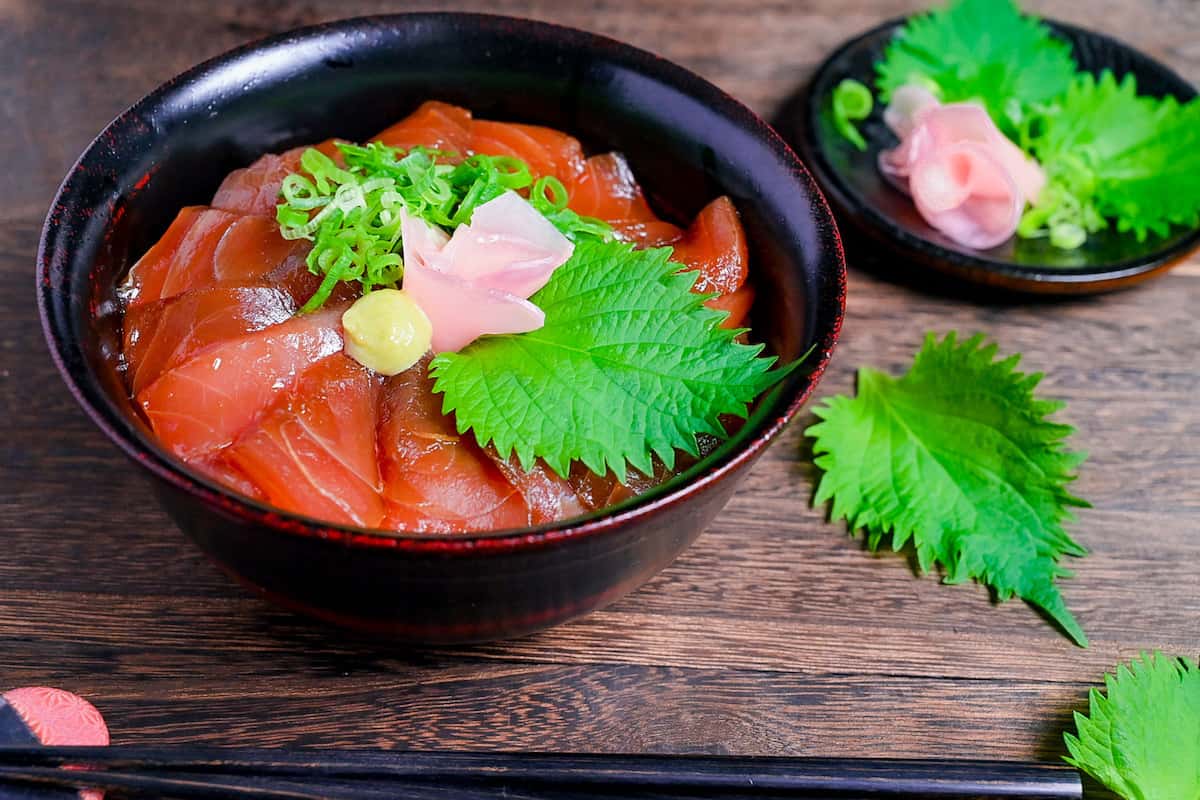 Tekkadon - vinegared rice topped with slices of fresh tuna sashimi, pickled ginger, wasabi, shiso and chopped spring onion in a black lacquerware bowl