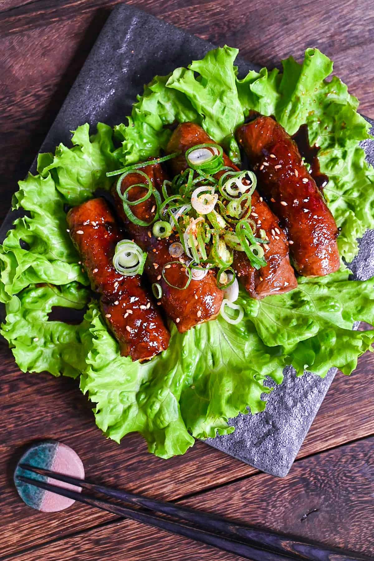 Beef Negimaki (Japanese Beef Scallion Rolls) served on a square black plate upon a bed of lettuce and sprinkled with sesame seeds