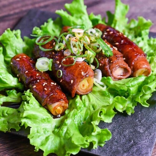 Beef Negimaki (Japanese Beef Scallion Rolls) served on a square black plate upon a bed of lettuce and sprinkled with sesame seeds