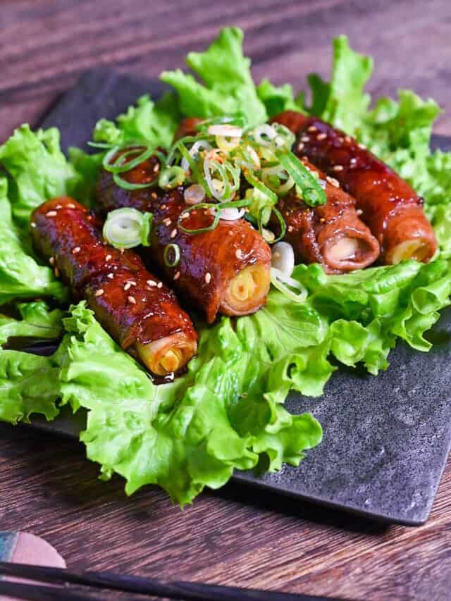 Beef Negimaki (Japanese Beef Scallion Rolls) served on a square black plate upon a bed of lettuce and sprinkled with sesame seeds