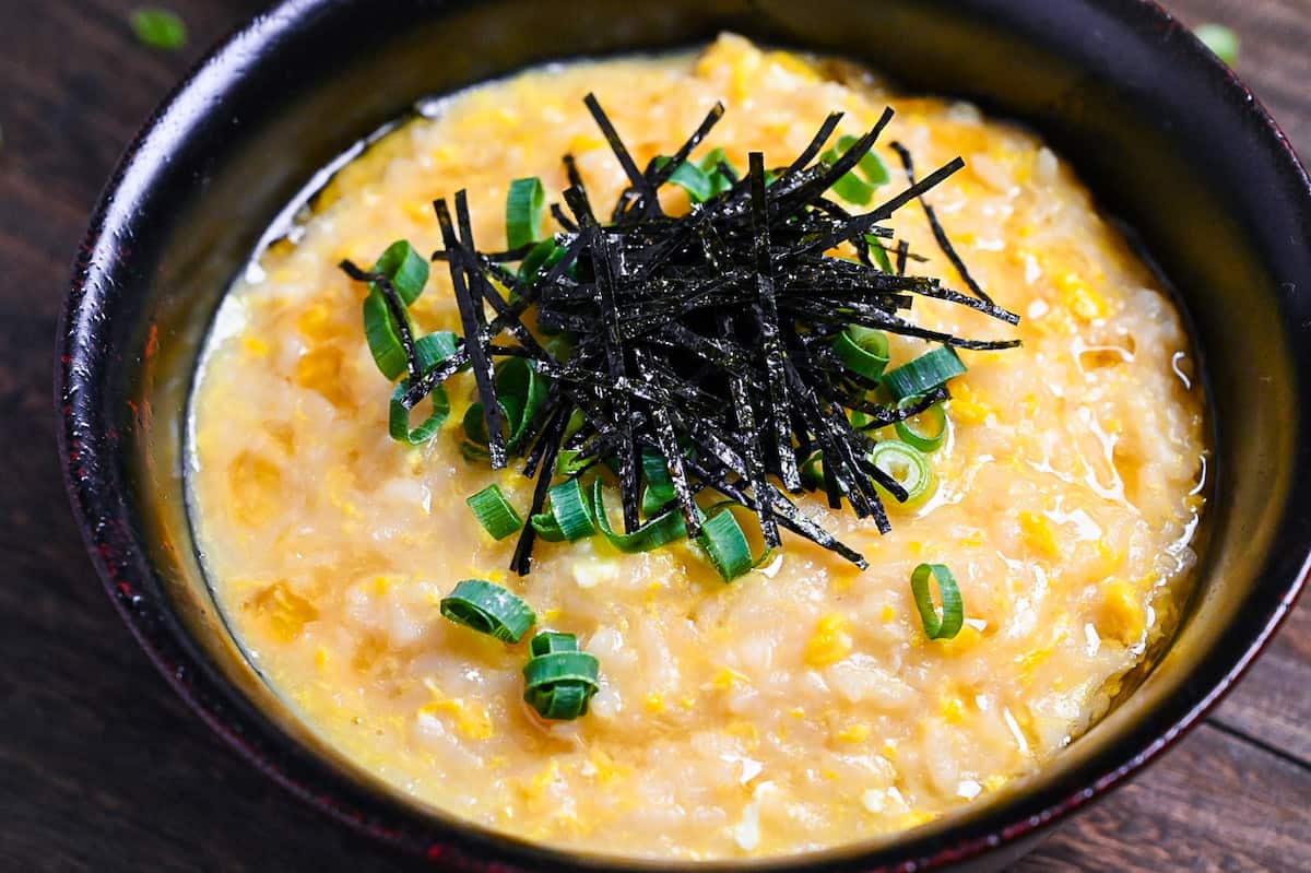 Okayu (Japanese Rice Porridge) with egg in a black lacquerware bowl sprinkled with spring onion and kizami nori