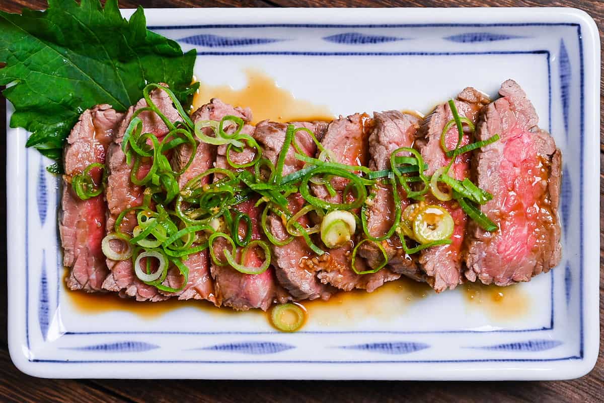 Beef tataki (Japanese seared beef fillet) on a white plate topped with homemade sauce and chopped green onions top down