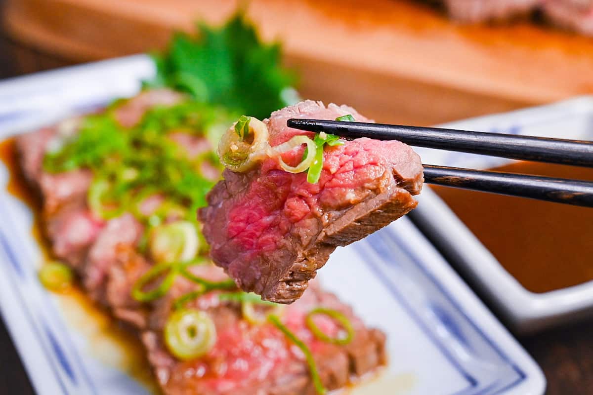 Beef tataki (Japanese seared beef fillet) on a white plate topped with homemade sauce and chopped green onions held with black chopsticks