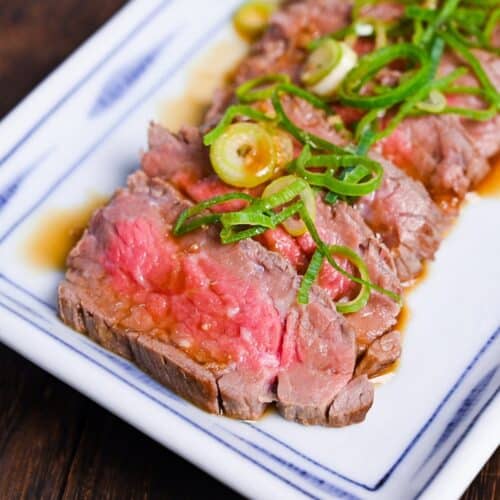 Beef tataki (Japanese seared beef fillet) on a white plate topped with homemade sauce and chopped green onions