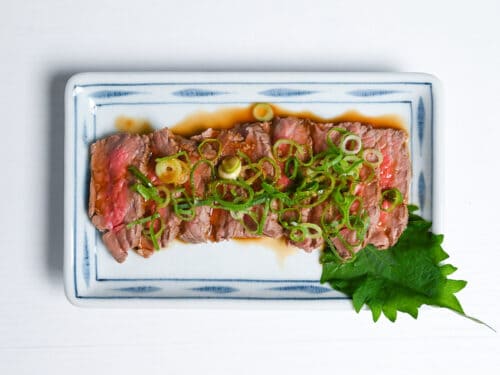 final beef tataki served on a white plate with sauce, finely chopped green onion and shiso leaf