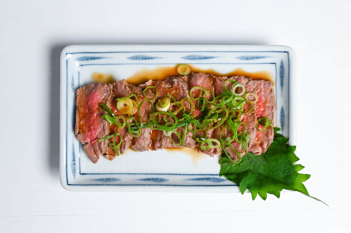 final beef tataki served on a white plate with sauce, finely chopped green onion and shiso leaf
