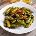 Stir fried edamame with garlic and oyster sauce glaze topped with dried chili and sesame seeds on a white plate
