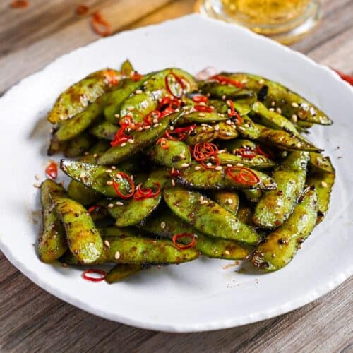 Stir fried edamame with garlic and oyster sauce glaze topped with dried chili and sesame seeds on a white plate